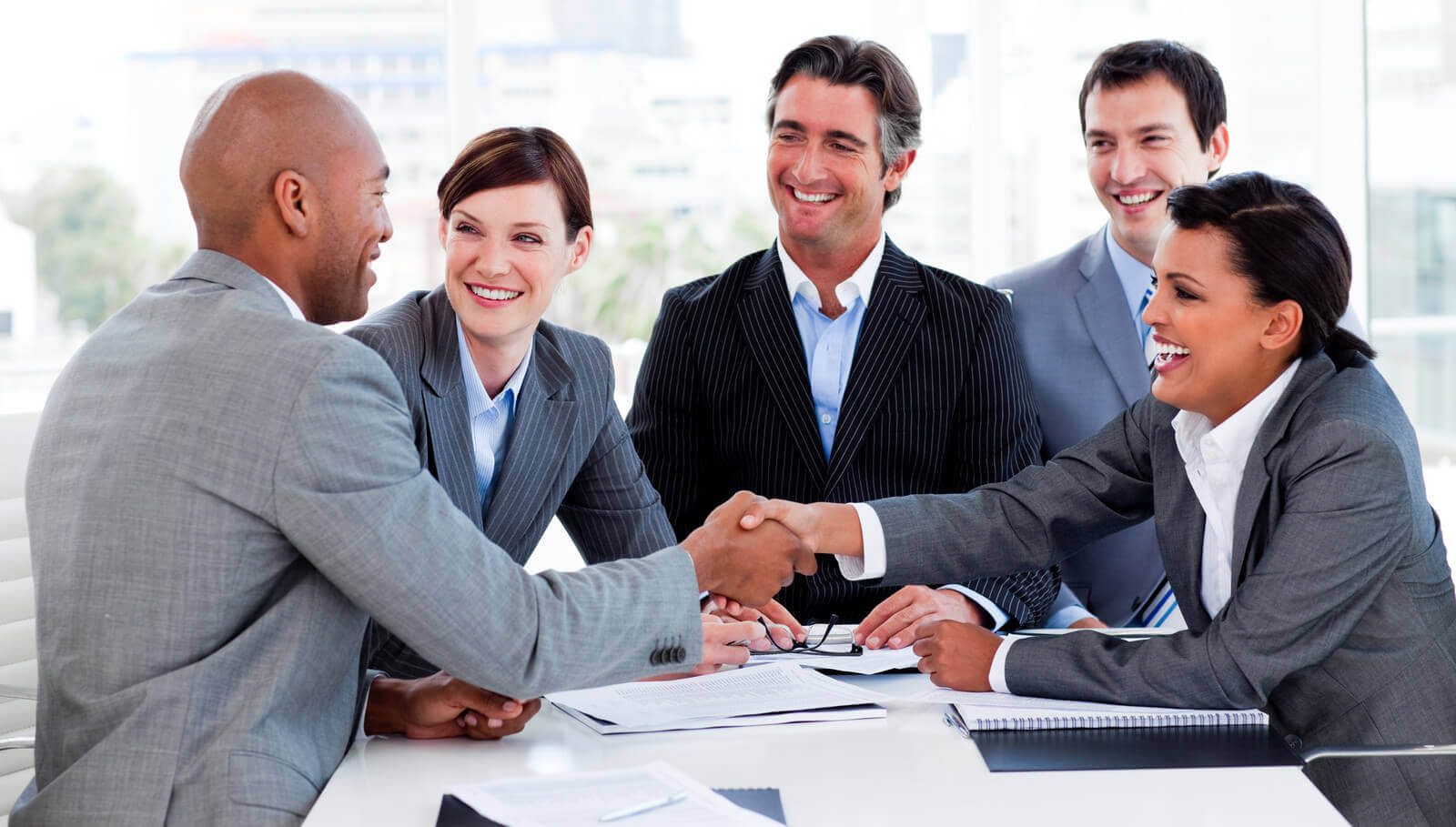 Five business women and men sitting at a table, two fo them shaking hands.