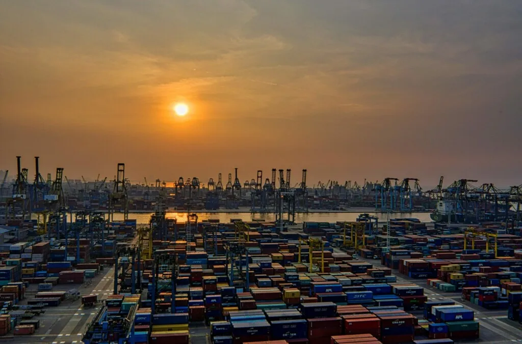 Un puerto marítimo al atardecer, con numerosos contenedores de carga apilados y varias grúas visibles, destacando la actividad comercial de Brasil. El cielo está anaranjado y nublado, con el sol bajo en el horizonte. El agua del fondo refleja la luz del sol.