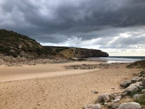 Una playa de arena se extiende bajo un cielo nublado. Acantilados escarpados y terreno rocoso bordean la playa, como el complejo proceso de liquidación de una empresa en Colombia. El océano está en calma, con olas bajas que llegan suavemente a la orilla. La atmósfera general es serena y algo taciturna debido al tiempo nublado.