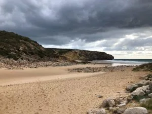 Una playa de arena se extiende bajo un cielo nublado. Acantilados escarpados y terreno rocoso bordean la playa, como el complejo proceso de liquidación de una empresa en Colombia. El océano está en calma, con olas bajas que llegan suavemente a la orilla. La atmósfera general es serena y algo taciturna debido al tiempo nublado.