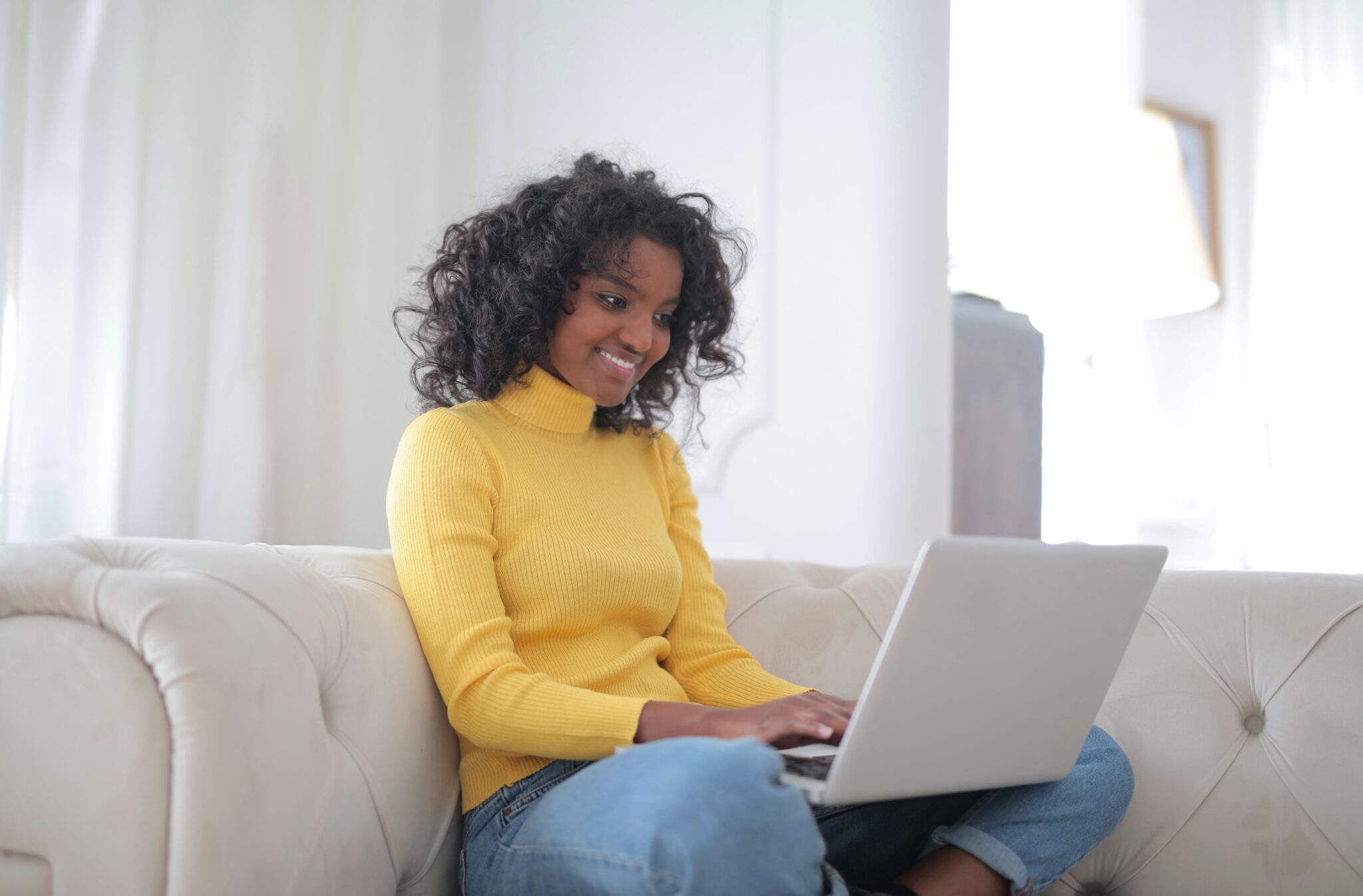 Uma pessoa vestindo um suéter amarelo e calça jeans está sentada em um sofá bege enquanto trabalha com um laptop. Ela tem cabelos cacheados e está sorrindo enquanto olha para a tela. O plano de fundo apresenta paredes brancas e uma janela clara.