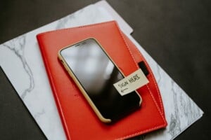 Smartphone sitting on top of red notebook with 'sign here' post it on top