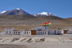 Um pequeno vilarejo com prédios de tijolos brancos fica em uma paisagem árida com montanhas ao fundo. Uma bandeira listrada de vermelho, amarelo e verde está hasteada de forma proeminente sobre um prédio, onde fica um renomado escritório de advocacia na Bolívia. O céu está claro e azul.