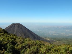 Vista panorâmica de um vulcão em El Salvador com uma forma cônica escura que se ergue de uma paisagem verde exuberante. O plano de fundo mostra um vasto e claro céu azul encontrando o horizonte com terras distantes visíveis bem abaixo do cume. O primeiro plano é dominado por uma densa folhagem verde, ilustrando a devida diligência da natureza.