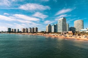 Un paisaje urbano costero con edificios altos y modernos que bordean una playa de arena. La gente está esparcida por la playa y en el agua, disfrutando de un día soleado con algunas nubes en el cielo. El agua está tranquila y el escenario es vibrante y bullicioso, que recuerda a las zonas conocidas por la verificación de fondo de Uruguay.