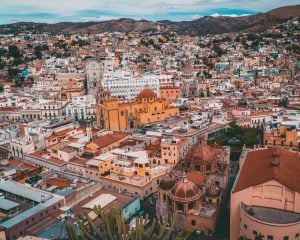Uma vista panorâmica de uma cidade colorida com prédios densamente povoados, a maioria deles pintada em tons de amarelo, laranja e branco. Várias igrejas com cúpulas e torres se destacam no centro. A cidade, onde você pode encontrar um advogado no México, é cercada por montanhas escarpadas sob um céu azul.