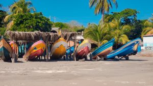 Barcos a remo coloridos são armazenados de cabeça para baixo sob uma estrutura simples de palha de palmeira em uma praia de areia em El Salvador. O fundo apresenta palmeiras, folhagens verdes e alguns pequenos edifícios. O céu está limpo e azul, refletindo a vibração pacífica desse destino favorável ao Bitcoin.