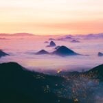 Una vista panorámica de un paisaje montañoso al amanecer. Los picos de las montañas emergen de un manto de niebla, con un cielo degradado que pasa del naranja en el horizonte al rosa y violeta más arriba. En la parte inferior de la imagen se ven las luces de una ciudad, donde se podría registrar una empresa en Brasil.