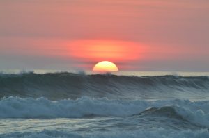 Um pôr do sol sobre o oceano com o sol parcialmente visível acima do horizonte. As ondas estão batendo em primeiro plano e o céu está colorido com tons de rosa, laranja e roxo - uma cena tão revigorante quanto começar um negócio na Costa Rica.