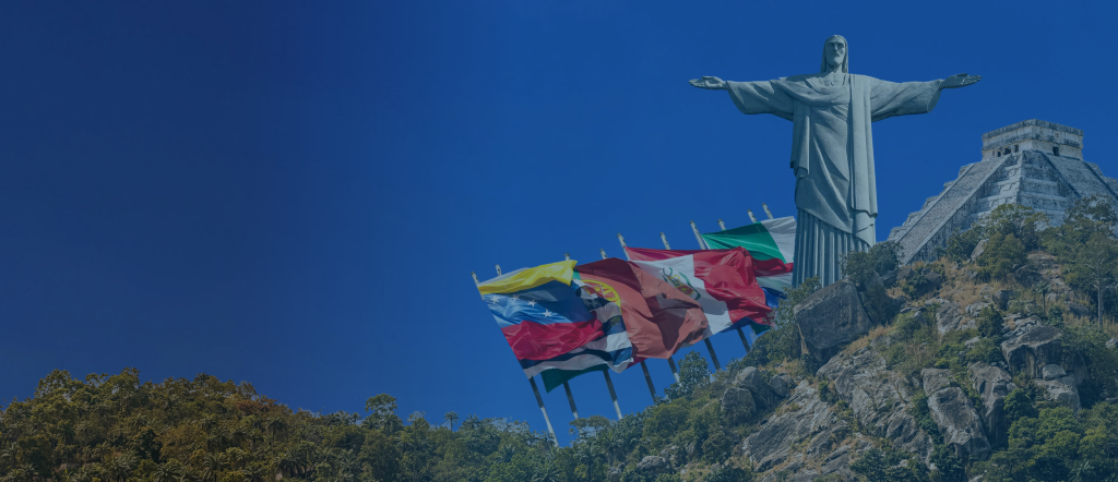 Header homepage BLH Una gran estatua del Cristo Redentor se alza sobre una colina rocosa, rodeada de vegetación. A la izquierda de la estatua se ven varias banderas nacionales, entre ellas las de Venezuela, Perú, Brasil y Argentina. El cielo azul despejado enmarca perfectamente este emblemático monumento en su pintoresco entorno.